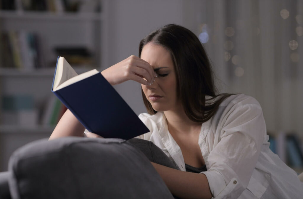 A young adult pinches the bridge of their nose to alleviate eye strain while attempting to read a book on a couch at home