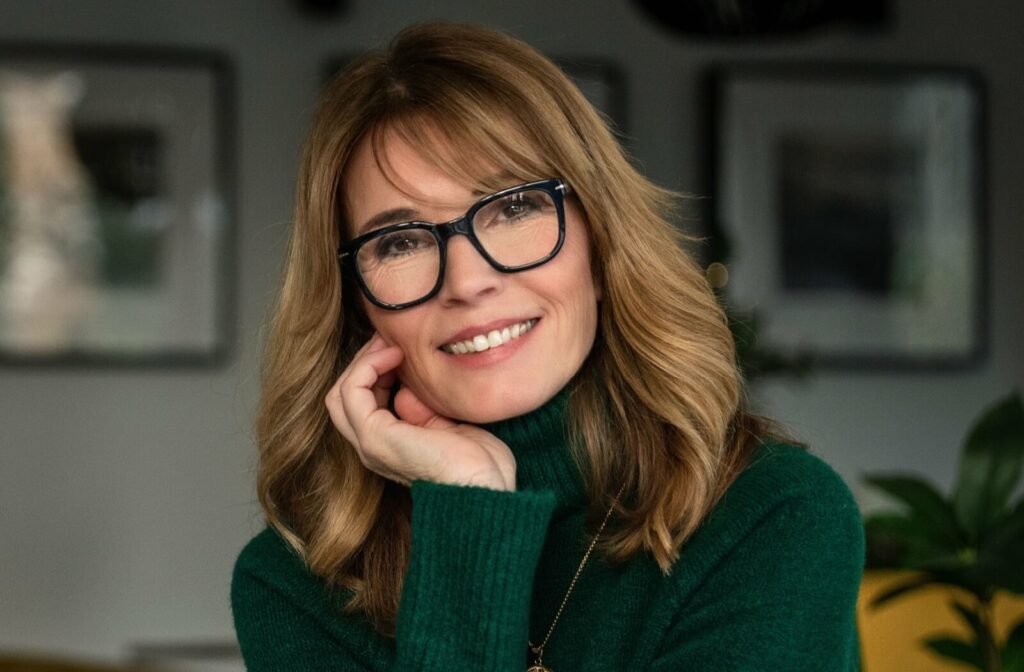 Person with long wavy hair smiling and wearing dark rectangular glasses and a green sweater indoors