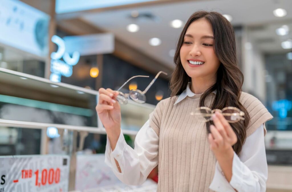 Person with long dark hair smiling while holding two pairs of eyeglasses in a retail eyewear store.