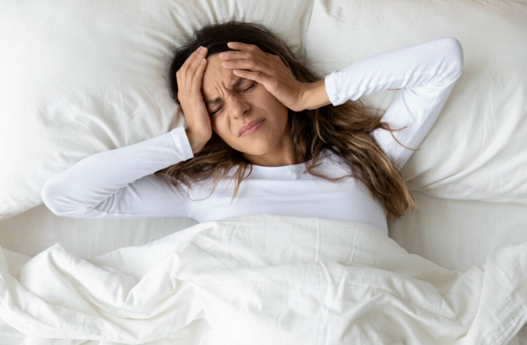 Woman lying in bed holding her head with both hands, looking uncomfortable as if experiencing pain or a headache.