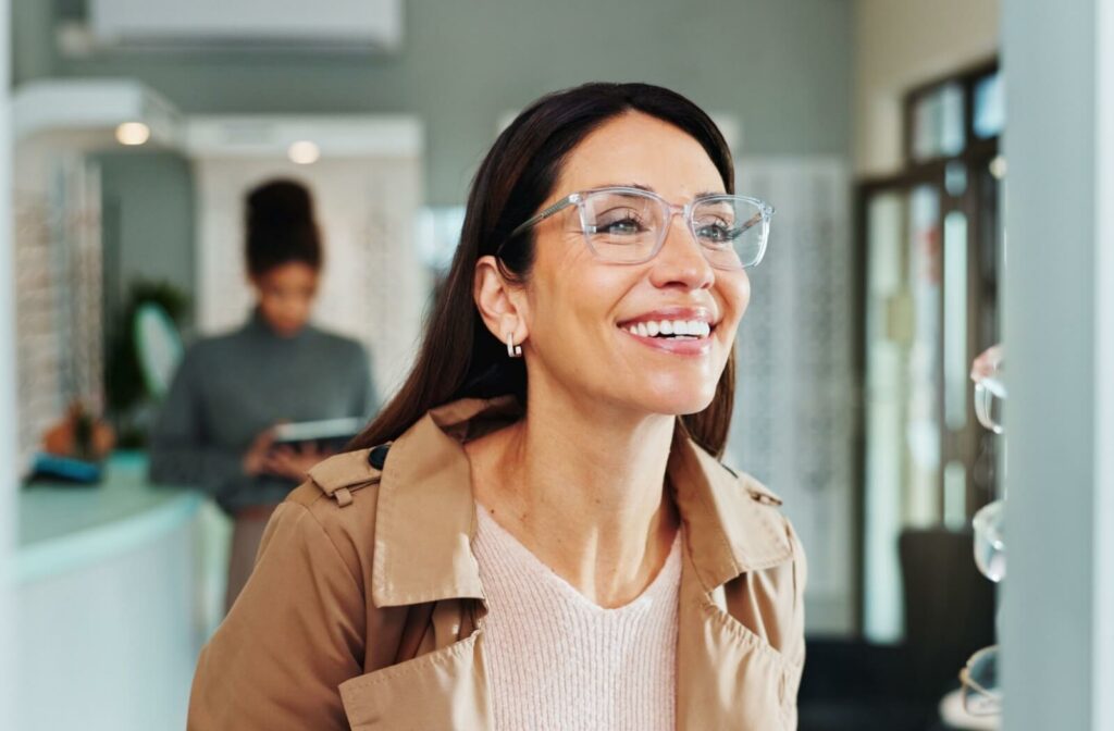 A person in a tan coat smiling while trying on clear-rimmed eyeglasses at an optical shop, showcasing 2026 eyewear trends.