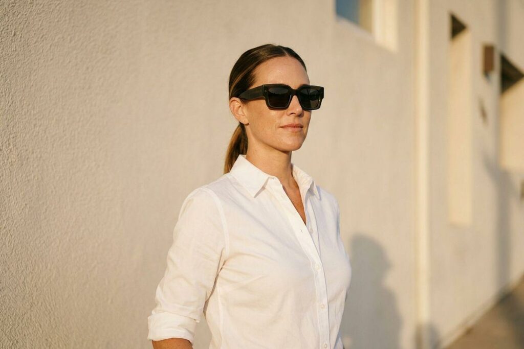 Person in black sunglasses and white button-up shirt standing outdoors near a light-colored wall in sunlight.